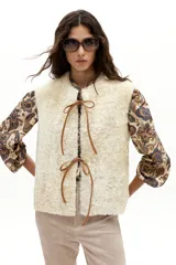 Chaleco reversible de gamulán de oveja en color natural, con corte holgado, cuello a la base y terminaciones al corte. Presenta tiras de cuero en el frente para atar.