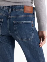 Jeans corte bootcut de tiro medio-bajo, confeccionado en denim azul con lavado desgastado y contrastes. Presenta cierre frontal con botón metálico y bolsillos laterales y traseros.