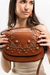 Cartera bandolera de cuero 100% natural color marrón, con estructura armada y tapa decorada con tachas plateadas colocadas a mano. Forrada en tela con bolsillo interno con cierre.
