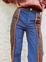 Pantalón de paño con silueta recta y levemente acampanada. Presenta un diseño de paneles combinados en azul marino y marrón, con detalles de vivos en cuero color naranja que recorren el largo de las piernas. Cuenta con cierre frontal y bolsillos laterales.