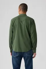 Camisa de hombre verde militar, estilo western, con cuello clásico, cierre de botones a presión blancos, dos bolsillos con solapa en el pecho y costuras decorativas en los hombros.