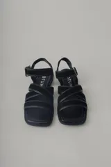 Sandalia plana de cuero negro, con diseño minimalista de tiras finas cruzadas sobre el empeine y una tira ajustable con hebilla metálica en el tobillo.