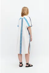 Túnica blanca de lino con corte oversized, escote en V, cierre con botones y mangas cortas. Presenta bordados geométricos azules en hombros y laterales.