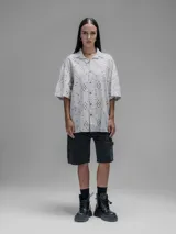 Camisa de manga corta color beige con cuello clásico y cierre de botones. Presenta un diseño de bordado calado con motivos florales y geométricos.