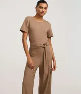 Mono de mujer color marrón topo, con escote redondo, mangas cortas y pantalón ancho. Incluye un cinturón ancho del mismo tejido que se anuda con una hebilla metálica redonda.