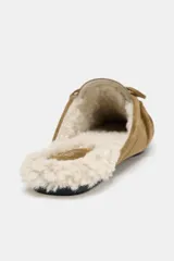 Mocasín destalonado tipo mule confeccionado en serraje color tostado, con detalle de fruncido y borlón en la parte delantera. Interior forrado en efecto borreguito y suela plana.