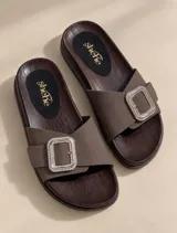 Sandalias planas color marrón con suela de aspecto amaderado y plantilla negra con logo dorado. Poseen una tira ancha de gamuza en el empeine decorada con una hebilla rectangular grande con pedrería brillante.