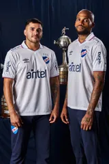 Camiseta de fútbol Umbro del Club Nacional de Football, modelo Home 2026. Diseño blanco con cuello en V, detalles en azul y rojo en el cuello y las mangas, y patrón en tejido jacquard. Incluye el logo de Umbro, el escudo del club y el sponsor antel en el pecho.