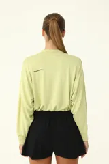 Buzo crop color verde claro, con cuello redondo y mangas largas holgadas. Presenta frunce en el ruedo mediante cordón elástico y tanca.