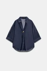 Camisa de denim azul oscuro con corte amplio, cuello solapa, escote en pico y mangas tres cuartos. Presenta un aplique metálico dorado en el centro y bajo asimétrico.