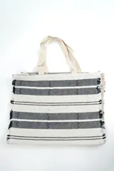 Bolso tipo tote bag tejido a mano, con diseño de rayas horizontales en color natural y azul marino, detalles geométricos en zigzag y flecos en los laterales. Cuenta con asas superiores de tela en tono crudo.