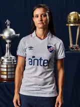 Camiseta de fútbol Umbro del Club Nacional de Football, modelo Home 2026 para mujer. Diseño en color blanco con textura jacquard que presenta siluetas de copas históricas en toda la prenda. Cuenta con cuello en V, detalles en azul y rojo en los bordes de las mangas y el cuello, logo de Umbro, escudo del club y sponsor Antel en el frente.