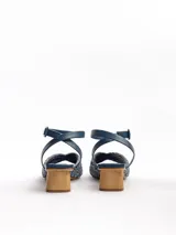 Sandalias de tacón medio en color azul, con capellada de rafia labrada en tonos azules y dorados, pulsera de cuero nobuck enlazada al tobillo con cierre de hebilla, y taco de madera natural lustrada de forma cilíndrica. Cuenta con puntera cuadrada.