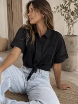 Camisa de manga corta color negro, con cuello camisero, cierre frontal con botones y detalles de encaje bordado en la parte delantera y en las mangas.