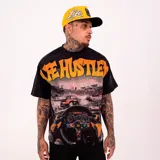Remera negra con diseño gráfico estilo racing que incluye una escena de automovilismo en el frente y la frase "Life Hustler" en letras grandes de color naranja con detalles brillantes. Tiene un corte oversize y mangas cortas.