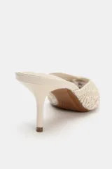 Zapato mule destalonado color crudo, con tacón fino alto, punta fina y cuerpo con bordados texturizados y apliques de cuentas.