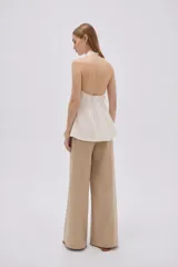 Blusa de algodón color beige con escote en V, cuello halter y cierre frontal con botones. Presenta un diseño ajustado en el torso con pinzas y pliegues en la parte inferior que generan una caída ligeramente acampanada.