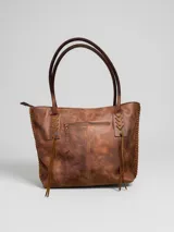 Cartera tipo tote bag de cuero vacuno marrón, con detalles de trenzado artesanal en los laterales y flecos colgantes decorativos. Cuenta con doble asa de hombro y cierre superior.