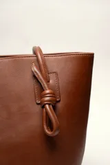 Cartera tote de cuero suave color marrón, con diseño amplio y líneas simples, con tiras de hombro decoradas con nudos artesanales y pequeño logo grabado en el centro.