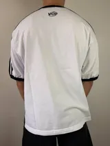 Remera de diseño estilo jersey deportivo con calce oversize, color blanco con mangas cortas negras y detalles de franjas blancas en los hombros. Cuenta con estampado frontal con texto y logotipos.