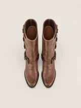 Bota de caña alta estilo biker, color marrón, con punta redondeada y taco cuadrado. Presenta múltiples tiras horizontales con hebillas metálicas y detalles de herrajes en el lateral. Confeccionada en cuero con forro vacuno y suela de madera.
