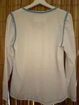Remera de manga larga blanca con escote en V y vivos en color celeste. Presenta un bolsillo frontal con estampado de rayas celestes y un diseño circular bordado en color amarillo. Las mangas tienen un ligero fruncido en los hombros.