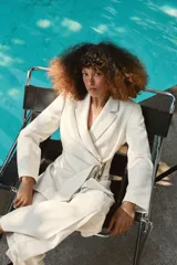 Blazer de corte estructurado en color beige, con cuello solapa y manga larga. Presenta un diseño cruzado con cierre mediante lazada del mismo tejido y un distintivo aplique metálico dorado en el frente.