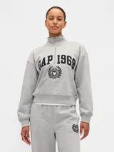 Buzo deportivo de color gris melange con medio cierre frontal y cuello alto. Presenta estampado frontal con el logo Gap 1969 y un emblema circular con un corazón. Posee mangas largas con puños elásticos y un corte cropped.