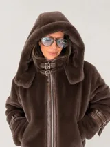 Campera de piel sintética color chocolate con capucha oversize. Presenta cierre frontal metálico, detalles en material engomado, cuello alto con doble hebilla y correas ajustables en los puños.