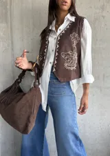 Chaleco color chocolate con bordados en hilo blanco en los bordes y frente. Presenta detalles de tachas metálicas en los hombros y cierre frontal.
