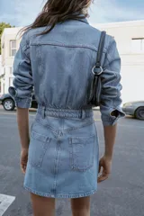 Vestido corto de denim azul claro, estilo camisa, con cuello solapa, escote pico, manga larga y cierre frontal de botones metálicos. Presenta bolsillos delanteros con tapa y bolsillos de plastrón en la espalda. La cintura está ajustada con elástico fruncido.