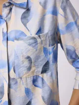 Camisa de manga larga con cuello clásico y cierre frontal de botones. Presenta un estampado de hojas en tonos azules sobre fondo claro y un bolsillo frontal en el pecho.