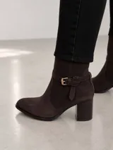 Bota de caña media confeccionada en cuero color marrón oscuro, con punta almendrada y taco cuadrado de 7 cm. Presenta una tira decorativa con hebilla metálica dorada en la zona del tobillo.