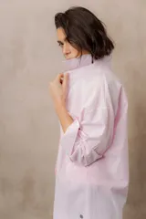 Camisa blanca de calce amplio, confeccionada en tejido liviano con sutiles rayas verticales, cuello clásico, mangas largas con hombros caídos y abotonadura frontal.