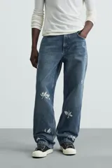 Jeans de corte relaxed fit en denim de algodón, color azul medio lavado. Presenta bordados florales en color blanco o crudo distribuidos en las piernas. Modelo de cinco bolsillos con cierre frontal de cremallera y botón.