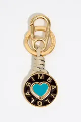 Llavero con mosquetón y argolla de metal dorado, con un cordón trenzado beige y negro. El dije principal es circular, esmaltado en negro, con un corazón turquesa en el centro y letras doradas alrededor que forman el nombre de la marca.