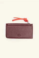 Monedero rectangular de cuero vacuno con textura granulada en color bordó. Cuenta con cierre superior metálico y un tirador de cuero en color naranja con nudo decorativo. Incluye un pequeño logo grabado en el centro.
