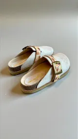 Sandalias de cuero blancas modelo Saki, estilo zueco, con hebilla metálica ajustable y detalle de diseño pintado a mano en la tira.