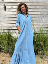 Vestido camisero largo de jean, con cierre frontal de botones, cuello camisero, mangas largas y detalle de volado en el ruedo.