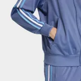 Campera deportiva Adidas Firebird azul con cierre frontal, cuello alto y tres rayas blancas y celestes en las mangas. Logo bordado en el pecho. Corte holgado y puños acanalados.