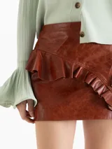 Minifalda de cuero ovino color marrón, de tiro medio-alto y silueta entallada. Presenta un recorte diagonal con un volado aplicado de forma asimétrica.