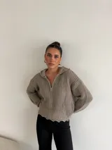 Buzo de punto grueso color gris, con cuello polo, cierre frontal parcial y capucha. Presenta un diseño de tejido acanalado con terminaciones desflecadas en el ruedo.
