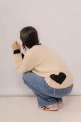 Buzo tejido a mano en lana merino, color crema con detalles en negro en el cuello, puños y un diseño de corazón en la parte inferior. Presenta un corte amplio y mangas largas.