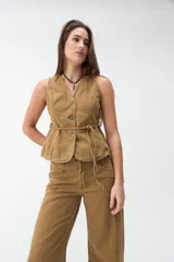 Chaleco de denim color beige, sin mangas, con escote en V, cierre frontal con botones y lazo ajustable en la cintura.