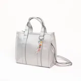 Cartera tipo tote bag color plateado con textura granulada, doble asa de mano y correa larga ajustable y extraíble. Incluye un colgante decorativo con cuentas de colores en una de las asas.