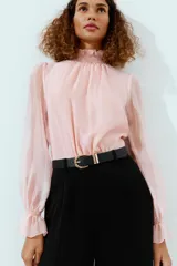 Mono largo con parte superior de blusa rosa de manga larga, cuello alto fruncido y puños con volantes. La parte inferior es un pantalón negro de pernera ancha con bolsillos delanteros y un cinturón negro con hebilla dorada.
