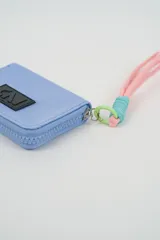 Billetera pequeña color turquesa con cierre perimetral y parche frontal negro con logo. Incluye una correa de muñeca de cordón rosado con detalle de hilo celeste.
