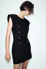 Vestido corto de estilo militar en color negro, con cuello redondo, hombreras y manga sisa. Presenta un diseño frontal con falsos botones dorados dispuestos en filas horizontales. Cierre lateral con cremallera oculta y abertura en la espalda con botón.