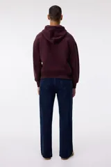 Campera con capucha de tejido pesado, color borgoña oscuro, con cierre frontal, bolsillos canguro y puños y bajo acanalados. Presenta un corte oversized y holgado.