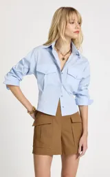 Camisa de corte cropped y entallado, color celeste, con cuello clásico, mangas largas con puños, cierre frontal de botones y dos bolsillos delanteros con solapa y botón.